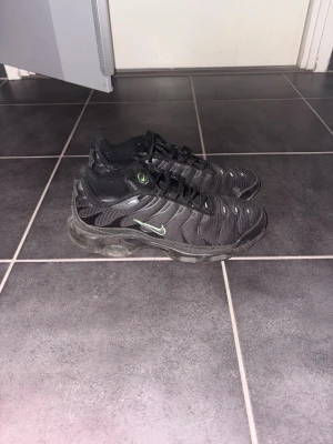 Nike tn Plus svarta sneakers - Svarta Nike tn Plus sneakers med mesh- och syntetöverdel, gröna detaljer och synlig Air-dämpning i sulan. Klassisk TN-logga på plösen och sidan. Snygg vågig design och låg profil som passar streetwear-stilen.