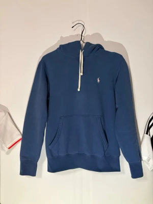 Blå hoodie från Polo Ralph Lauren - Snygg blå hoodie från Polo Ralph Lauren med klassisk huva och vita dragsnören. Tröjan har en känguruficka framtill och den ikoniska lilla loggan broderad på bröstet. Tillverkad i mjukt bomullsmaterial som är skönt att ha på sig.