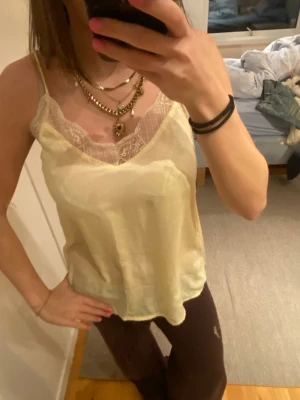 Ljusgult spets linne  - Säljer ett ljusglt spets linne med tunna axelband och v-ringning. Toppen har fina spetsdetaljer längs urringningen och är tillverkad i ett glansigt material som ger en lyxig känsla. Perfekt att styla med smycken för en trendig look.