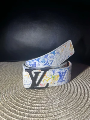 Louis Vuitton vitt bälte med färgstänk - Säljer ett vitt bälte från Louis Vuitton med färgstänk i blått, gult och orange samt klassiskt LV-monogram. Bältet har en stor svartfärgad LV-logga som spänne och är gjort i präglat läder. Perfekt för dig som vill sticka ut med en exklusiv accessoar.