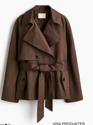 Slutsåld oanvänd brun trenchcoat med knytskärp med lappen kvar  - Stilren brun trenchcoat från H&M med bred krage, dubbelknäppning och knytskärp i midjan. Jackan har två fickor med knappdetaljer och är tillverkad i ett mjukt bomullsliknande material. Perfekt för lager på lager och en klassisk look.