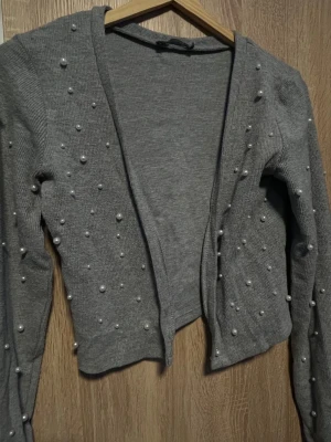 Grå kofta med pärldetaljer från Zara  - Snygg grå ribbstickad kofta med dekorativa pärlor över hela plagget. Koftan har öppen front och långa ärmar, vilket ger en trendig och stilren look. Perfekt för dig som vill ha något extra till din outfit. Använt 3 gånger 😊🫶🏽