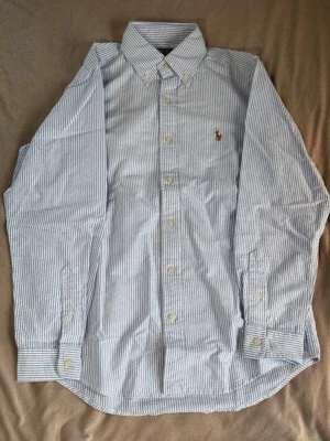 Randig skjorta från Polo Ralph Lauren - Klassisk blå- och vitrandig skjorta från Polo Ralph Lauren i bomull. Skjortan har button-down krage, långa ärmar och knappar framtill. Broderad logga på bröstet och relaxed passform. Perfekt till jeans eller chinos. Sitter mer en S