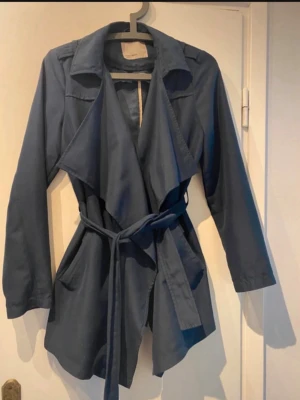 Mörkblå trenchcoat från Vero Moda - Stilren mörkblå trenchcoat från Vero Moda med skärp i midjan och stora slag. Jackan har långa ärmar, två fickor och ett mjukt, följsamt tyg som ger en snygg siluett. Perfekt för dig som vill ha en klassisk men modern look.