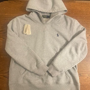 Grå hoodie från Polo Ralph Lauren - Snygg grå hoodie från Polo Ralph Lauren med klassisk huva och känguruficka. Tröjan har det ikoniska lilla mörkblå logobroderiet på bröstet och ribbade muddar vid ärmar och nederkant. Tillverkad i mjuk bomullsblandning för extra komfort.