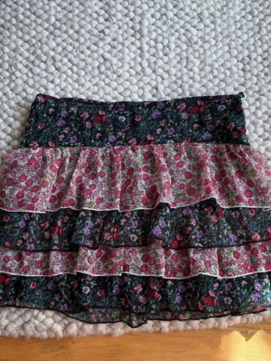 Blommig volangkjol från 365 Sunshine - Supersöt kort blommig kjol från 365 Sunshine med flera lager volanger. Kjolen har svart botten med små blommor i rosa, rött, lila och grönt. Perfekt för dig som gillar romantisk och lekfull stil. Tunn och luftig, passar till många outfits! Passar en Xs till M