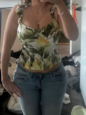 Linnetopp med citronmönster - Säljer en söt linnetopp med volangaxelband och citronmönster i grönt och gult på vit botten. Toppen har en croppad passform och är perfekt för sommardagar. Materialet känns lätt och luftigt, och axelbanden har fina volangdetaljer. den täcker min navel och jag är 160 cm 