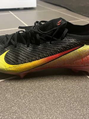 Nike Mercurial fotbollsskor svart/gul, vinicus jr limited edition - Säljer ett par Nike Mercurial fotbollsskor i svart med stora gula och röda detaljer. Skorna har snörning, låg profil och är tillverkade i syntetmaterial för bästa grepp och känsla på planen. Perfekta för dig som vill sticka ut på fotbollsplanen.