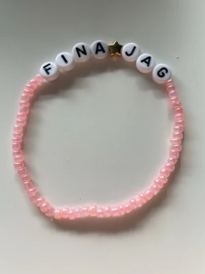 Pärlarmband med text och stjärna - Sött armband med ljusrosa pärlor och vita bokstavspärlor som stavar 'FINA JAG'. Mellan orden sitter en liten guldfärgad stjärnpärla. Perfekt accessoar för att ge en personlig touch till din stil.