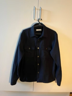 Mörkblå overshirt från Mango - Stilren mörkblå overshirt i Ull från Mango med klassisk krage, två bröstfickor med lock och knappar framtill. Tillverkad i ett kraftigare tyg som ger en snygg och avslappnad look. Perfekt att bära över en t-shirt eller hoodie. Mitt pris: 349. Nypris: 999.