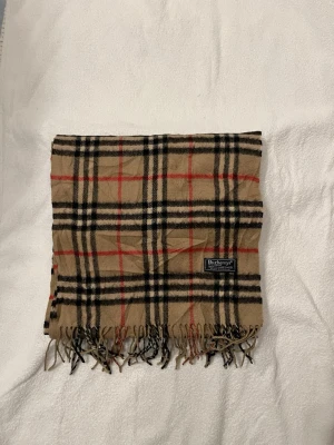 Burberry scarf - Klassisk beige halsduk från Burberry med det ikoniska rutmönstret i svart, vitt och rött. Halsduken har fransar i båda ändar och en mjuk känsla, perfekt för att lyfta vilken outfit som helst.