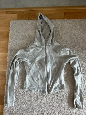Ljusgrå hoodie med dragkedja - Snygg ljusgrå hoodie med dragkedja framtill och figurnära passform. Hoodien har långa ärmar och en klassisk huva. Tillverkad i mjukt bomullsmaterial som känns skönt mot huden. Perfekt till jeans eller leggings för en avslappnad look. Storlek L 