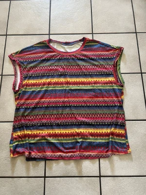 Flerfärgad ärmlös randig topp storlek 1XL - Säljer en färgglad ärmlös topp med randigt mönster i rött, gult, blått, grönt och svart. Toppen har rund halsringning och är tillverkad i ett mjukt syntetmaterial. Perfekt för dig som gillar statement-plagg och vill sticka ut.