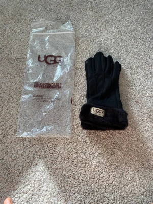 Svarta pälsfodrade handskar från UGG - Stilrena svarta handskar från UGG med mjuk pälsdetalj vid mudden och broderad logotyp. Perfekta för kalla dagar när du vill hålla händerna varma och samtidigt se snygg ut. Klassisk design som passar till det mesta.