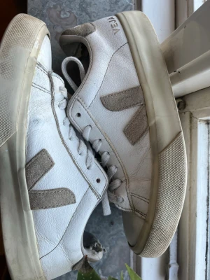 Vita Veja sneakers med beige detaljer - Säljer ett par klassiska vita Veja sneakers med beige mockadetaljer och logga på sidan. Skorna har lågt skaft, vita snören och en robust sula i gummi. Perfekta för dig som gillar stilrena och tidlösa sneakers med en clean look. Dem är lite risiga i hälen men utöver det är dem jätte snygga och sköna 