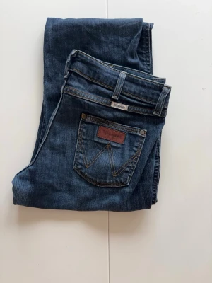 Mörkblå jeans från Wrangler midjemått 39 - Snygga mörkblå jeans från Wrangler med klassisk femficksdesign och kontrastsömmar. Jeansen har normal passform och är tillverkade i bomull för en bekväm känsla. Perfekta till din vardagsstil!  storlek i läng L 32 har Obs har lite litet där nere men inte alls mycket 