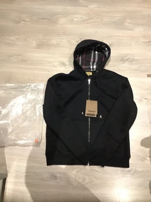 Svart hoodie från Burberry - Säljer en svart zip up från Burberry med dragkedja och klassiskt rutigt foder i huvan. Hoodien har fickor framtill och justerbar dragsko i huvan. Perfekt för dig som gillar stilrena plagg med exklusiva detaljer.