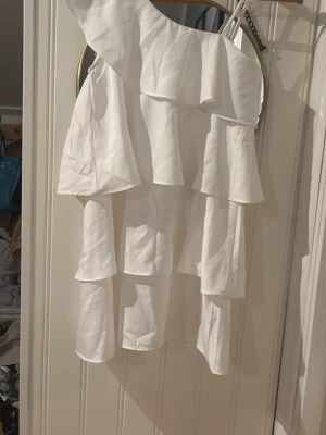 Vit oneshoulder volangklänning - Säljer en vit oneshoulder klänning med flera lager volanger. Klänningen har en asymmetrisk axel och är ärmlös, vilket ger en trendig och ungdomlig look. Perfekt för sommarens alla tillfällen.