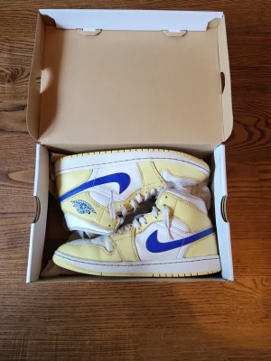 Nike Air Jordan 1 Mid gul/blå - Säljer ett par Nike Air Jordan 1 Mid sneakers i fräscha färger. Skorna är gula och vita med blå swoosh och detaljer. Klassisk Jordan-logga på sidan och hög siluett. Tillverkade i läder med vita skosnören och matchande sula. Perfekta för dig som gillar streetwear.