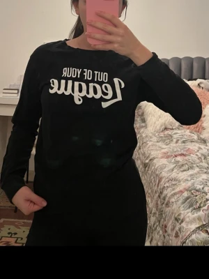 Svart långärmad t-shirt med tryck - Svart långärmad t-shirt med vit text på bröstet där det står 'Out of your league' i en cool font. T-shirten har rund halsringning och en avslappnad passform. Perfekt för dig som gillar statement-plagg och enkel stil.