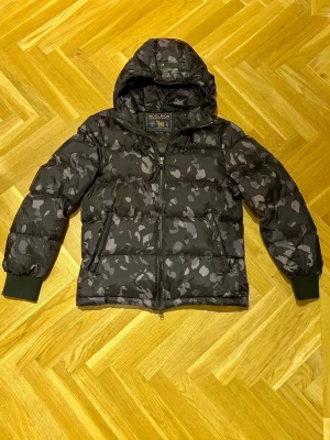 Woolrich puffer jacka camomönster - Säljer en svart och grå camomönstrad pufferjacka från Woolrich med huva och dragkedja framtill. Perfekt orent runt då den inte är så tjock som man tror. Storlek S, mycket bra skick.