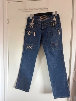Blå jeans med broderi från CBB - Säljer ett par blå jeans från CBJ med raka ben och coola broderade detaljer på fickorna. Jeansen har dekorativa band och X-detaljer i beige samt flera fickor både fram och bak. Perfekta för dig som vill ha en unik och trendig look. Köpta på vinted