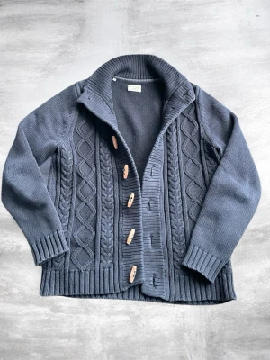 Mörkgrå stickad kofta med träknappar - Säljer en mörkgrå stickad kofta med grovt kabelstickat mönster och snygga träknappar. Koftan har hög krage, ribbade muddar och nederkant samt en avslappnad passform. Perfekt för dig som gillar klassisk men ändå trendig stil.