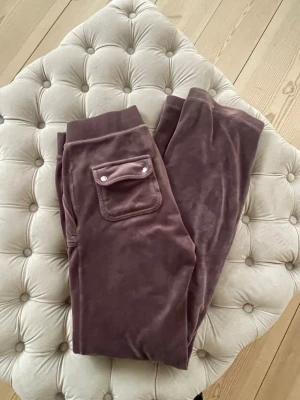 Bruna Juicy Couture  - Säljer ett par mjuka och sköna mjukisbyxor i brun velour. Byxorna har bred mudd i midjan och en ficka med lock och knappar på sidan. 