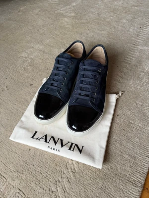 Mörkblå sneakers från Lanvin - Snygga mörkblå sneakers från Lanvin med ovandel i mocka och lackad svart tå. Skorna har vita sulor och klassisk låg profil. Perfekta för dig som vill ha en stilren och exklusiv look med en twist av lyx. Storlek UK11 motsvarar ungefär 45-46.