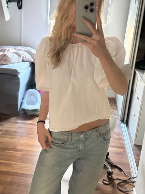 Vit puffärmad blus med knyt bak - Säljer en söt vit blus med puffärmar och tre knytband i ryggen. Blusen har en luftig passform och är tillverkad i bomull, perfekt för en fräsch och trendig look. Passar till både jeans och kjol för en avslappnad stil.