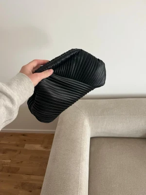Svart plisserad kuvertväska/clutch - Snygg svart clutch med plisserad design och mjuk struktur. Väskan har en modern, stilren look och är tillverkad i ett material som liknar konstläder. Använd en gång.