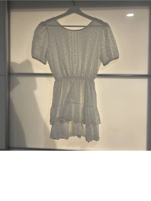 Vit kort spetsklänning med volang - Superfin vit kortklänning från Lucilia med broderade spetsdetaljer och volang i kjolen. Klänningen har korta puffärmar, markerad midja och v-ringning i ryggen med en knap. Perfekt för sommarens! Kläningen är totalt 76 cm från axlarna och ner (bortsett från markerade midja) ! Kläning går ej att köpa eller hitta annars💗☀️