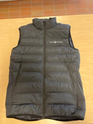 Svart dunväst från Sail Racing - Snygg svart dunväst från Sail Racing med hög krage och två dragkedjeförsedda fickor framtill. Västen har quiltad design och logga på bröstet. Perfekt för lager-på-lager och håller dig varm under kyliga dagar.