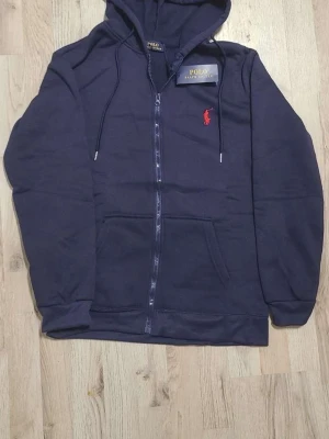 Mörkblå hoodie från Polo Ralph Lauren - Mörkblå hoodie med dragkedja från Polo Ralph Lauren. Klassisk design med huva och dragsnören, samt röd broderad logga på bröstet. Mjuk bomullsmix och kängurufickor framtill. Perfekt för en avslappnad och stilren look.