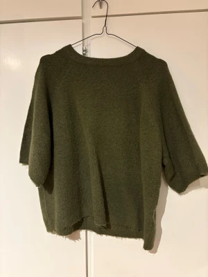 Olivgrön stickad tröja med kort ärm - En enkel och stilren olivgrön stickad tröja med korta ärmar och rund hals. Tröjan har en lös passform och ribbstickad kant vid halsen. Perfekt för lager på lager och passar till både jeans och kjol.