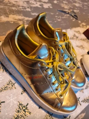 Guldiga Adidas Stan Smith sneakers - Som Nya knappt använda Unika Adidas Stan Smith sneakers i metallic guld med vita sulor och gula skosnören. Klassisk låg modell med perforerade detaljer på sidan och Stan Smith-tryck i rött. Tillverkade i syntetmaterial med bekväm innersula.