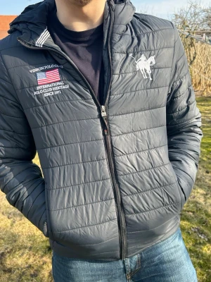 Svart pufferjacka från Vinson Polo Club - Snygg svart/blåaktig pufferjacka från Vinson Polo Club med broderad logga och amerikansk flagga på bröstet. Jackan har huva, dragkedja framtill och diskreta horisontella sömmar. Perfekt för kalla dagar och riktigt stilren look.
