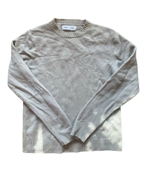 Ljusgrå stickad tröja från Samsoe Samsoe - Ljusgrå stickad tröja från Samsoe Samsoe med rund halsringning och ribbade muddar vid ärmslut och nederkant. Tröjan är enkel och stilren, perfekt för lager-på-lager och har långa ärmar. Materialet är mjukt och tunt, vilket gör den skön att bära. Det förekommer några små hål i tröjan.