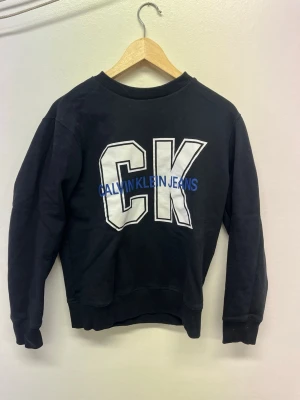Svart sweatshirt från Calvin Klein - Svart sweatshirt med stora vita bokstäver 'CK' och blå text 'Calvin Klein Jeans' framtill. Tröjan har rund halsringning, långa ärmar och ribbade muddar vid ärmslut och nederkant. Perfekt för en avslappnad och trendig look.
