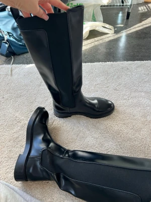 Svarta höga stövlar med elastiska paneler - Skitsnygga svarta stövlarmed högt skaft och breda elastiska paneler på sidorna. Kan egentligen användas alla årstider. Snygga till kjol/shorts på sommaren/våren men även på hösten/vintern. Köpte på sellpy men aldrig använt de då jag aldrig kännt att de passat min stil. ser som nya ut och är väldigt glansiga och snygga. Syns egentligen bara på undersidan att de är använda. 