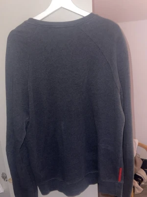 Prada sweatshirt - Mörkgrå sweatshirt från Prada med klassisk passform och rund halsringning. Tröjan har en diskret röd Prada-logga på ärmen och är tillverkad i mjukt material men Perfekt för en stilren och avslappnad look.