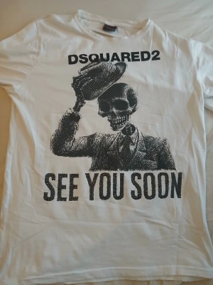 Vit DSQUARED2 t-shirt med tryck - Säljer en vit t-shirt från DSQUARED2 med stort svart tryck av en skelettgubbe i kostym och texten 'SEE YOU SOON' framtill. Texten är dekorerad med små pärlor för extra detalj.