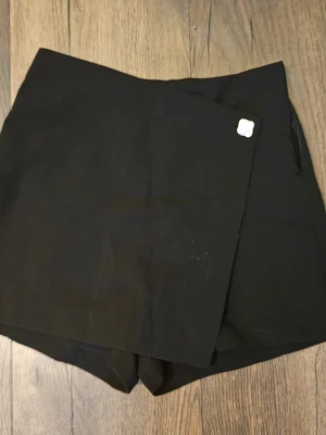 Svart skort med omlottlook - Stilren svart skort med omlott-design framtill och dragkedja i sidan. Klassisk och enkel modell som passar till många olika stilar. Tillverkad i ett mjukt tyg som ger en bekväm känsla. Perfekt för dig som vill ha shortskomfort men kjolens look.