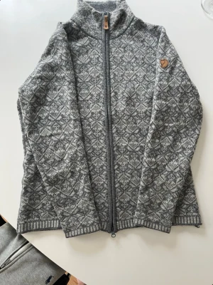 Grå mönstrad ullkofta från Fjällräven - Säljer en gråmelerad ullkofta från Fjällräven med hel dragkedja och hög krage. Fjällrävens klassiska logga på ärmen. Perfekt för kalla dagar och riktigt mysig. Nypris 2499kr! Köpt på Naturkompanet! Finns inga spår av noppor eller av användning. 