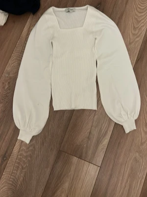 Vit ribbad topp från AWARE - Säljer en vit ribbad topp från AWARE med fyrkantig ringning och ballongärmar. Toppen har breda muddar vid ärmsluten och är croppad i modellen. Perfekt för dig som gillar trendiga detaljer och stilren design.