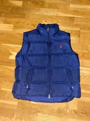 Mörkblå dunväst från Ralph Lauren - Snygg mörkblå dunväst från Ralph Lauren med hög krage och dragkedja framtill. Västen har två fickor med dragkedja och den klassiska röda Polo-loggan broderad på bröstet. Perfekt för lager på lager och riktigt skön att bära. Materialet är glansigt och quiltat.