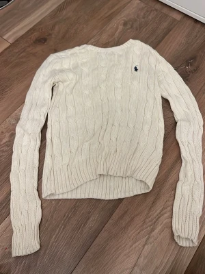 Vit kabelstickad tröja Polo Ralph Lauren - Kabelstickad tröja i mjuk pima bomull från Polo Ralph Lauren. Klassisk vit färg med rund halsringning och lång ärm. Liten mörkblå logga broderad på bröstet. Perfekt till jeans eller kjol för en clean och tidlös look.