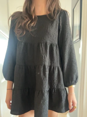 Svart kort klänning med puffärm - Säljer en svart kort klänning med puffiga långärmar och struktur i tyget. Klänningen har en lös passform och volangdetaljer i flera lager som ger en trendig look. Perfekt för dig som gillar en avslappnad men ändå stilsäker outfit.