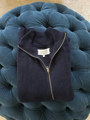 Mörkblå ziptröja från Maison Margiela - Snygg mörkblå ribbstickad ziptröja från Maison Margiela. Tröjan har hög krage och dragkedja framtill, vilket ger en clean och modern look. Perfekt för lager på lager och enkel att styla till olika outfits.