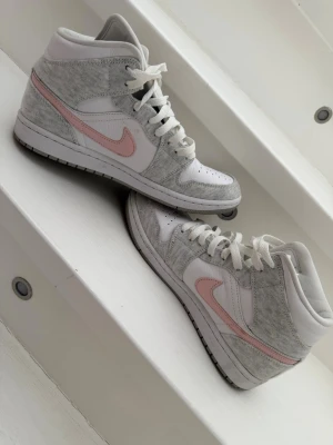 Nike Air Jordan 1 Mid grå/rosa - Snygga Nike Air Jordan 1 Mid sneakers i vitt och ljusgrått med rosa swoosh. Ovandelen är i en mix av mocka och läder, med klassiska perforeringar på tån och vita skosnören. Air Jordan-logga på sidan ger extra streetkänsla.
